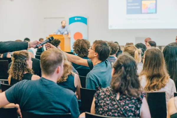 A room full of people, seen from the back, one person is being handed a microphone. At the far end of the room, someone is standing at a lectern.
